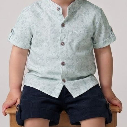 Child wearing a light green shirt and navy shorts sitting on a wooden stool against a plain background