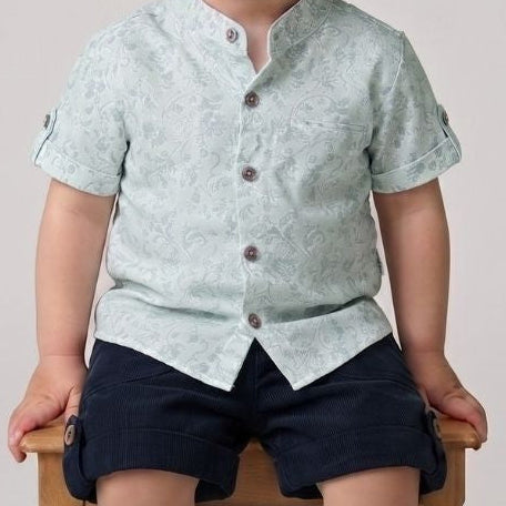 Child wearing a light green shirt and navy shorts sitting on a wooden stool against a plain background