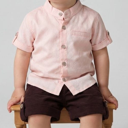 Child wearing a light pink shirt and dark shorts sitting on a wooden stool against a plain background.